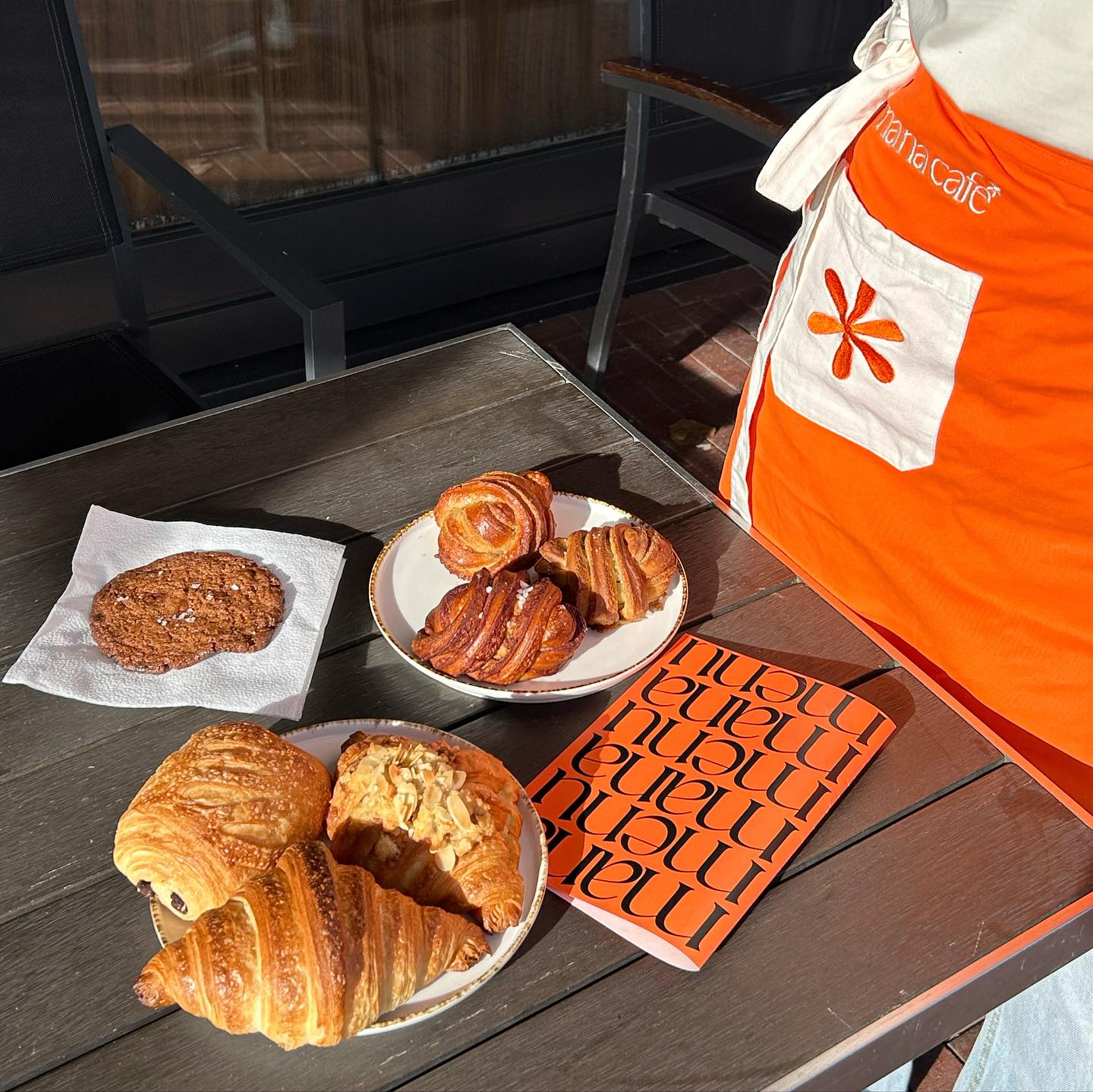 Croissants, kaneel-kardemombollen en een koekje op borden naast een oranje Mana-schort en menukaartje